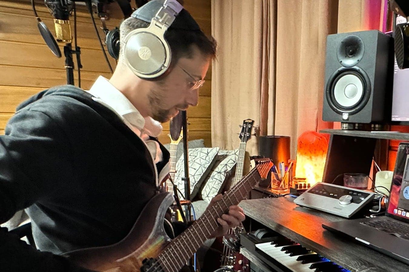 Moshe Siegel in headphones playing guitar at the studio desk, beside a monitor speaker and keyboard
